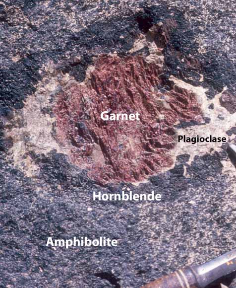 photograph of garnets in outcrop showing rim of plagioclase and hornblende in amphibolite host rock; hammer for scale