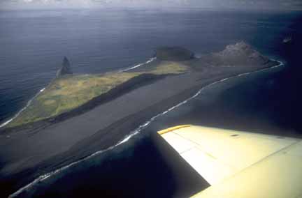 Photograph from air of black sandy beach and dark lava domes on volcanic island, grass-covered and bare rock in middleground, surrounded by ocean, plane wing in foreground