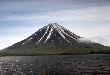 Photograph from boat of symmetrical, dark, volcanic peak with snow patches and green lower slopes; choppy dark blue ocean in foreground, sun glinting off water