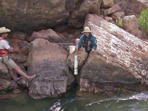photo of two workers wearing life belts standing on the rocks as the water flows past them.  One person holds an instrument about half a meter long