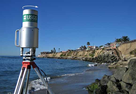 photo of instrument about the size of an oatmeal canister sitting on a tripod on the beach.  Cliffs in the background