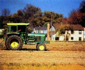 A color photograph of a John Deere tractor plowing in front of a housing development.