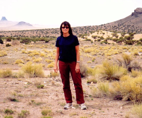 Photograph of USGS geologist Hiza Redsteer standing in front of aridlands in Arizona showing sand dunes and sage in foreground and monumental buttes in background. Dark red-colored buttes in the background.