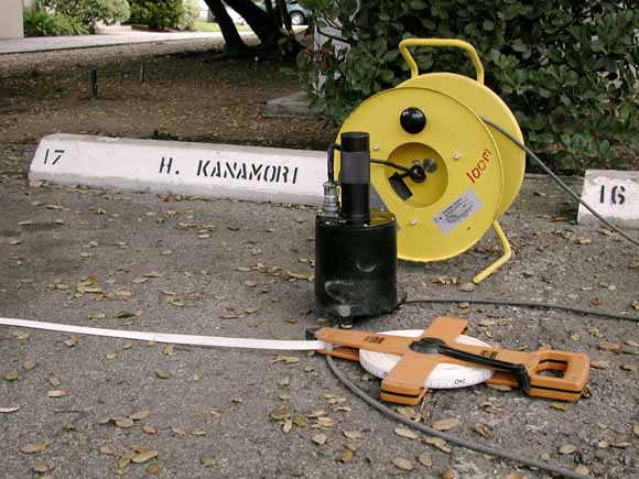 Photograph of seismometer, coil of data cable, and very long measuring tape.  The seismometer is a calendar and accessories about half a meter tall