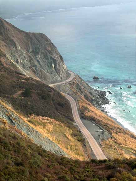Photo of narrow, winding Highway 1 taken from way up on a hill showing a big landslide scar on the uphill side and the surf crashing against the cliffs on the downhill side