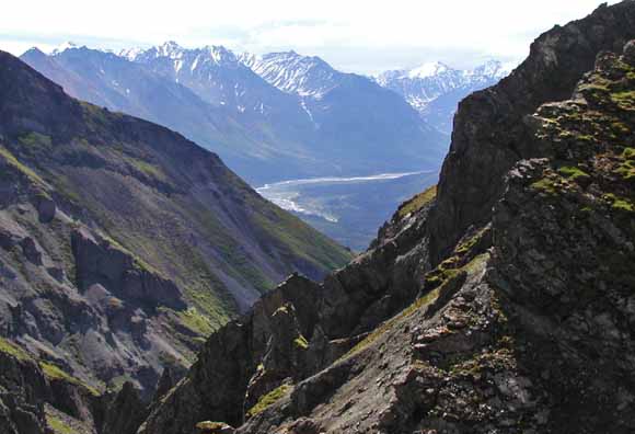 Photo looking down from between steep canyon walls at a river valley.  Snow-capped mountains are present in the background.