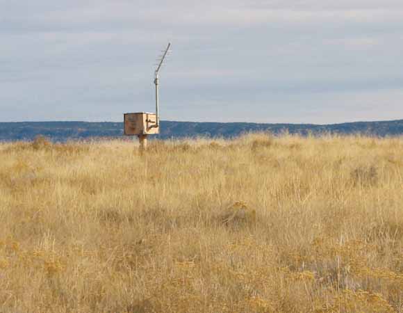 photo of dark-colored mesa in background with well and antenna in dry vegetation in foreground