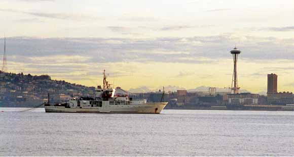 A picture showing ship described in caption below. The Seattle cityscape is in the background, including the Space Needle on the right. The airgun array and multichannel seismic streamer are deployed off the stern. 