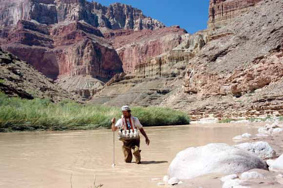 photo of man sampling water while wading in muddy river; cliffs in background