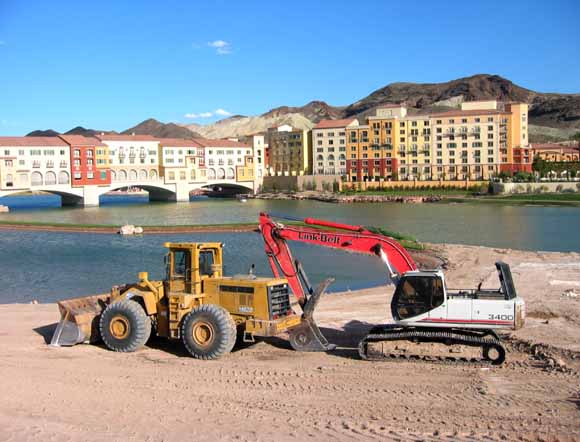 photo of dry hills in background, earthmoving equipment in the foreground, and a large artificial lagoon in the middleground with development around it