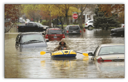 Man using in an inflatable boat to row down a flooded street that has several abandoned
                        cars.