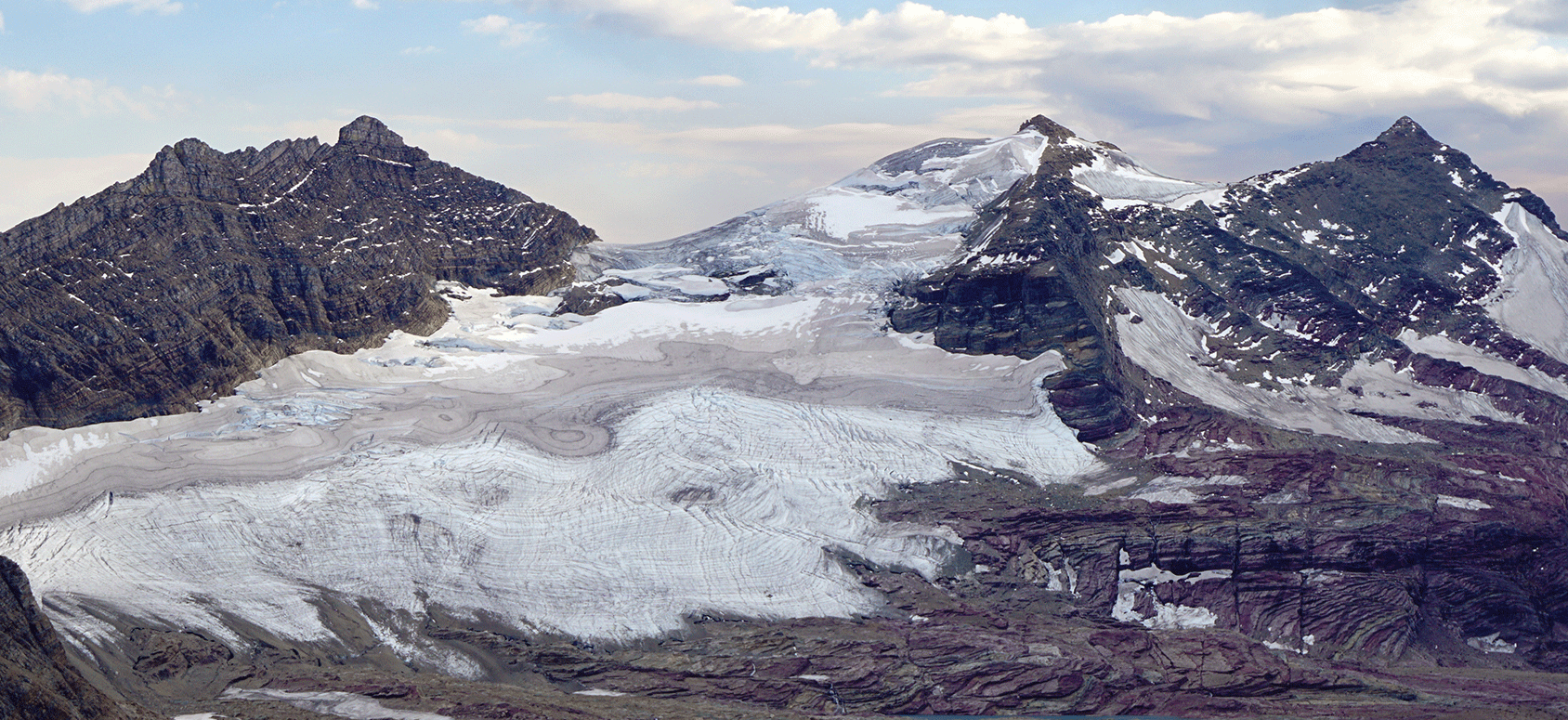 A glacier lies between dark rocky mountains. The top of the glacier has white and
                     gray areas.