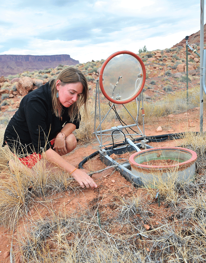 A woman bends over desert soil and vegetation next to equipment. A rocky cliff is
                           in the distance.