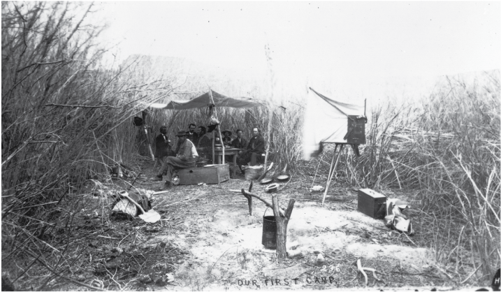 Makeshift tent-canopy covers eight people seated around a table at a campsite amidst
dense shrubbery.