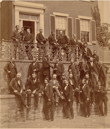Black and white image of four rows of men in suits, some sitting, most standing, near
a street edge outside of a building.