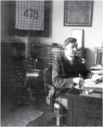A man seated in a chair at a desk holding a paper. The flag with the number “478”
is in the background.
