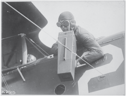 Man in an airplane wearing goggles and flight cap leans over the side of a plane with
the large camera.