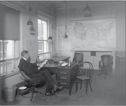 Man seated at a desk in an office reading from a sheet of paper. A wall-map of the
United States is opposite him.
