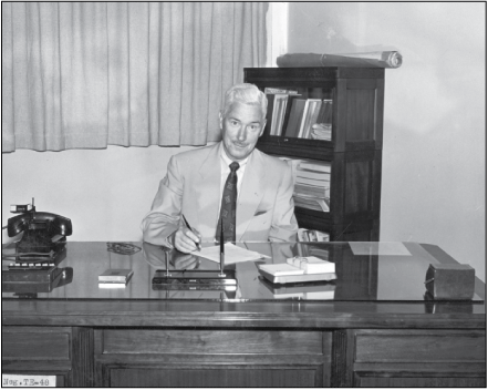 Smiling man in an office seated behind a desk writing on a piece of paper with a pen.