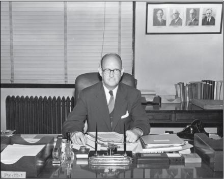 Man wearing glasses in an office seated behind a desk with several manuscripts in
front of him.