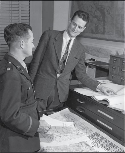 A man in a U.S. Army in front of a map, and a man in a suit pointing at a page in
a manuscript. The men are talking.