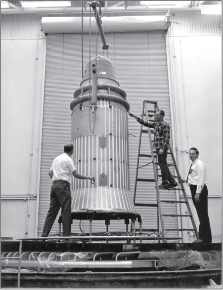 Three men around a tall satellite on a platform in front of a large roll-up bay door.
One man stands on a ladder.