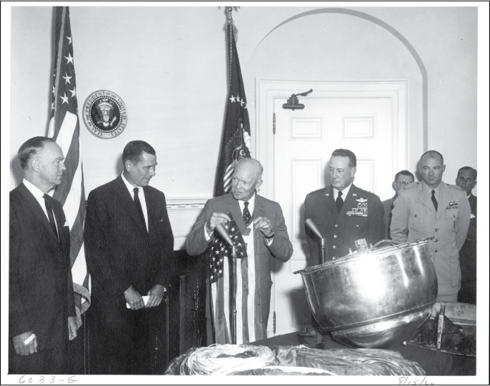 Seven men standing before a closed door in an office. A Corona satellite capsule sits
on a table.