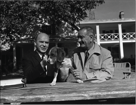 Black and white image of two men seated at a picnic table and a house in the background.
The President holds a beagle.