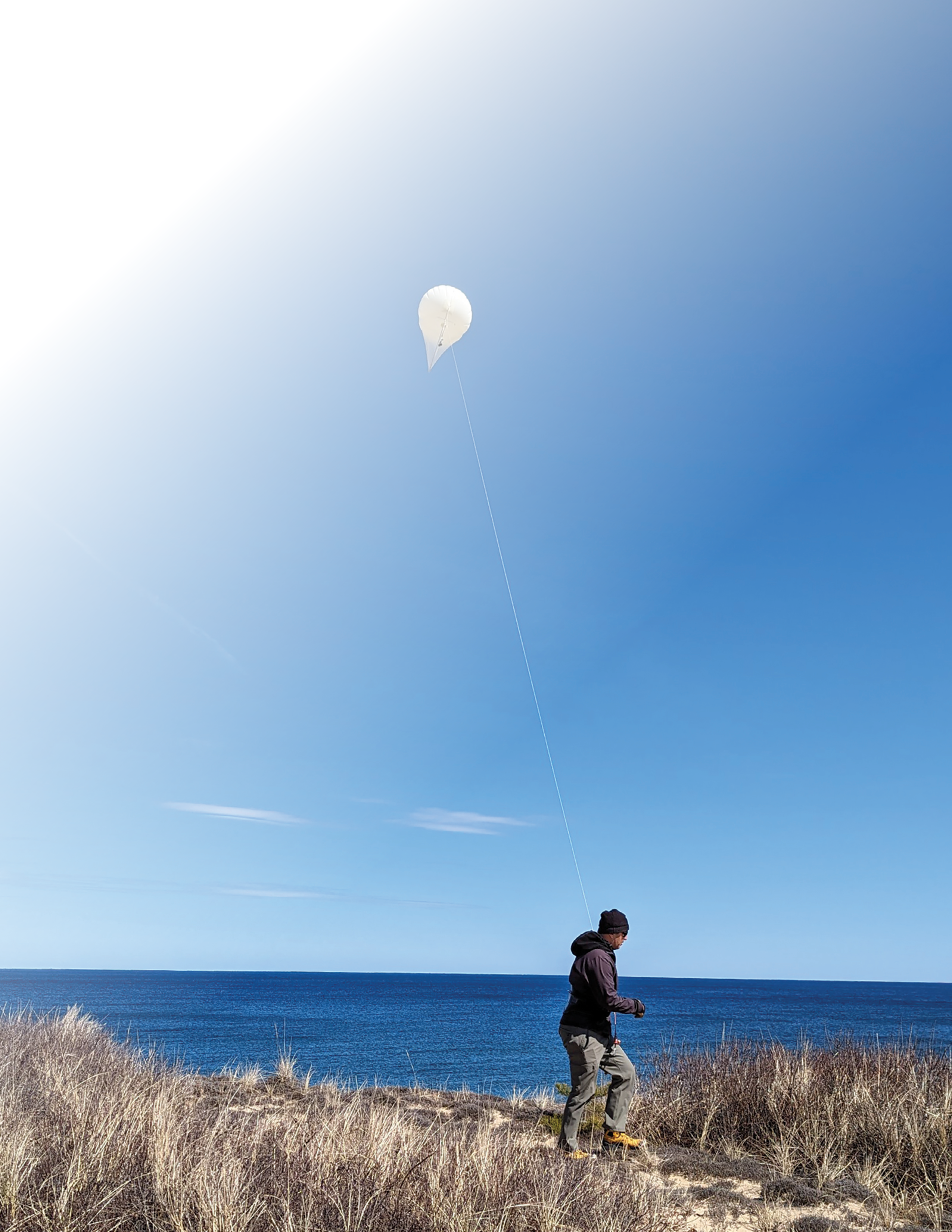 A scientist stands or walks on dunes partly covered in vegetation while holding a
cord. The helikite floating at the end of the cord appears mostly round and has a
sail with a pointed tip.