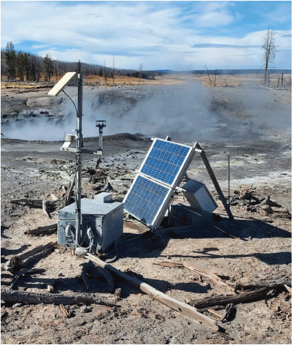 10. A short pole with equipment next to a metal box and solar panel on a barren ground
overlooking a steaming water pool. Forested mountains in the distance under partly
cloudy sky.