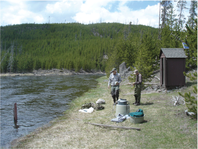 05. Two people with monitoring equipment near a small brown shed on the grassy banks
of a river. Tree-covered hill in the distance under a mostly cloudy sky.