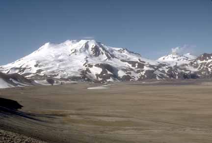 Photograph of snow-covered volcano, one steaming, in background and flat valley floor in foreground