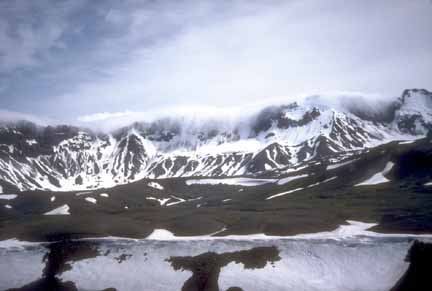 Photograph from within caldera of snow-covered rim in background and clouds pouring over from outside