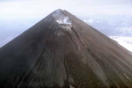 Photograph from air of dark, debris-covered, steaming, conical volcanic peak