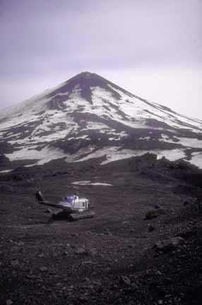 Photograph of partially snow-covered, conical volcano, helicopter on floats in foreground