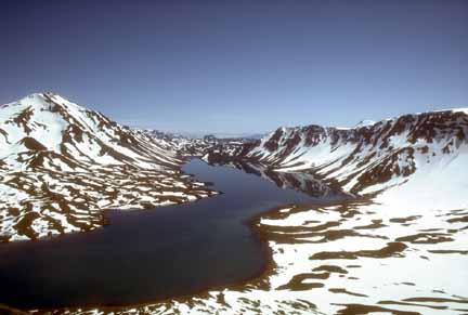 Photograph of blue, crescent-shaped lake inside snow-covered caldera, volcanic cone at left and caldera rim at right