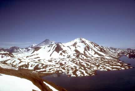 Photograph of snow-covered volcanic peaks with blue lake in foreground