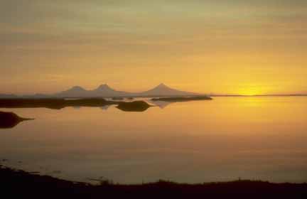Photograph of golden sunset reflecting off flat ocean, three silhouetted volcanoes on horizon
