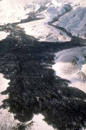 Photograph from air of slightly steaming, dark lava flow winding through snow-covered hills