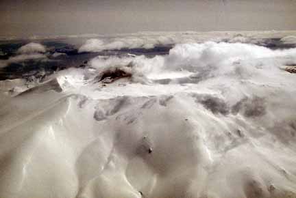Photograph from air of snow-covered volcano and dark, steaming cone in crater; clouds over ocean in background