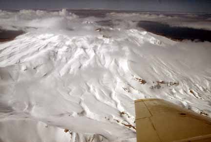 Photograph from air of snow-covered volcano, steam mixed with clouds around crater and plane wing in foreground