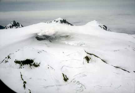 Photograph from air of snow-and ice-covered volcanic crater, peaks of rock and clouds in distance, steam coming from small crater in ice surrounded by dark ash