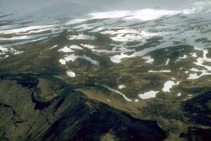 Photograph from air of snow partially filling volcanic craters and snowy landscape in distance, steep seacliffs in foreground
