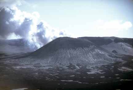 Photograph from air of steaming, ash-covered cone, caldera rim in background