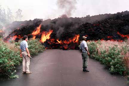 photo 010.  Photo of two men standing on road as lava, red-hot underneath and gray crusted-over on top, tumbles toward them