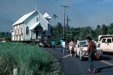 photo 067.  Photo of little white church up on wheels being towed down the middle of the road by the tractor from an eighteen wheeler.  Many workers walking along with it