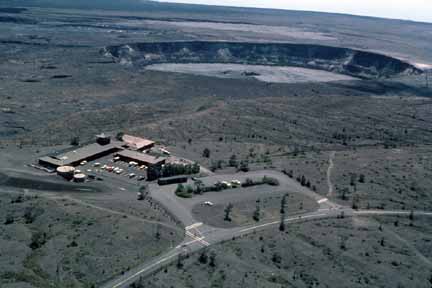 photo 100.  Low-elevation oblique aerial photo of complex of several buildings with parking lot in foreground, crater in background 