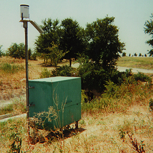 A. Photograph showing equipment shelter and solar panel for undeveloped site in Plano, Tex.; the rain gage is mounted just above the solar panel.