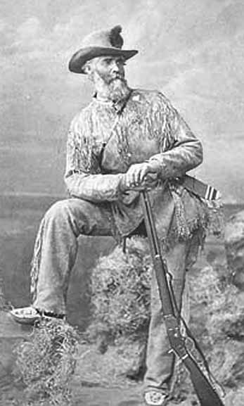 old black-and-white photograph of gray-bearded old man dressed in buckskins leaning on his lever-action Winchester Model 73 carbine (rifle).  Backdrop of clouds and valley and foreground of rocks and brush appear to be studio props.