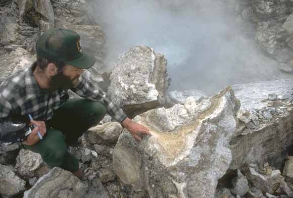 Recent color photograph of black-bearded young man dressed in park-service uniform leaning on broken boulder.  Backdrop of steam  and foreground of rocks are real.
